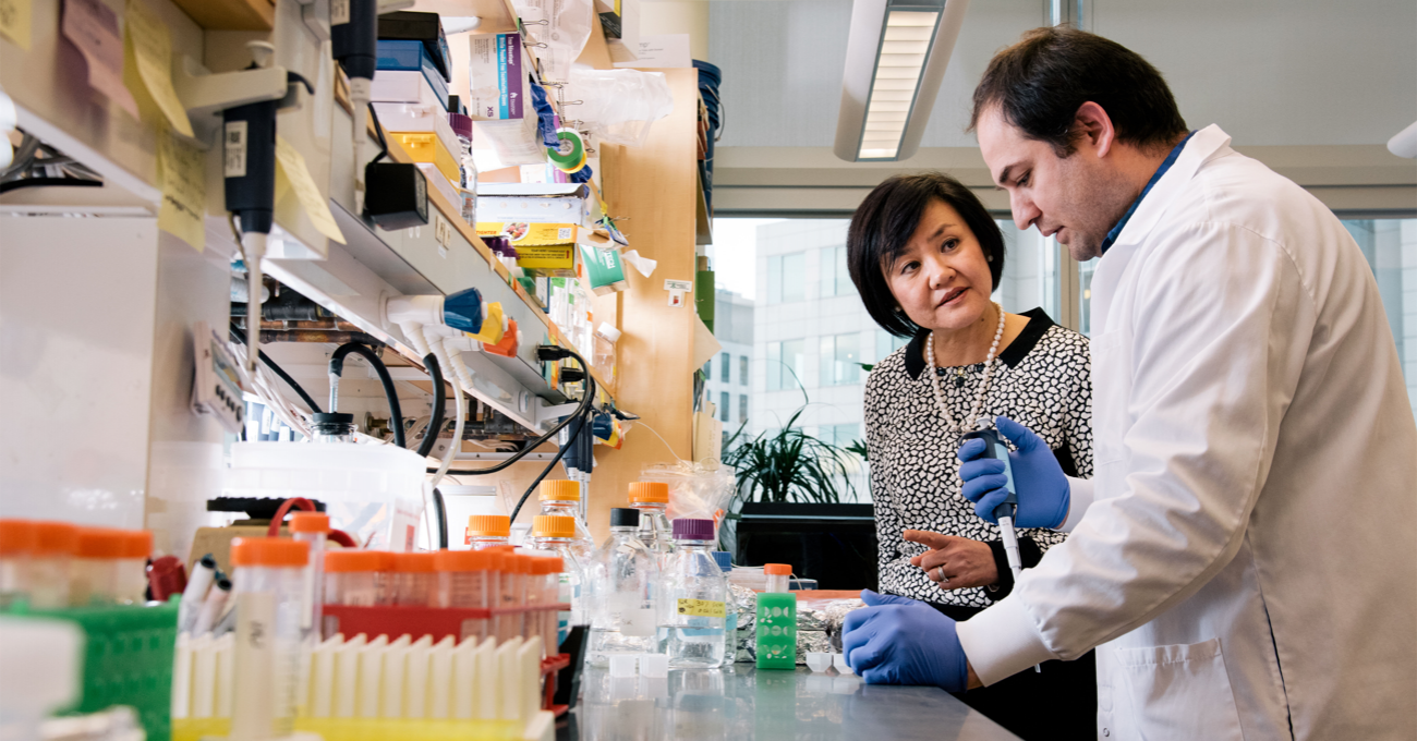Two neuroscientists stand at a lab bench. One, wearig gloves and a white coat, works with a pipette