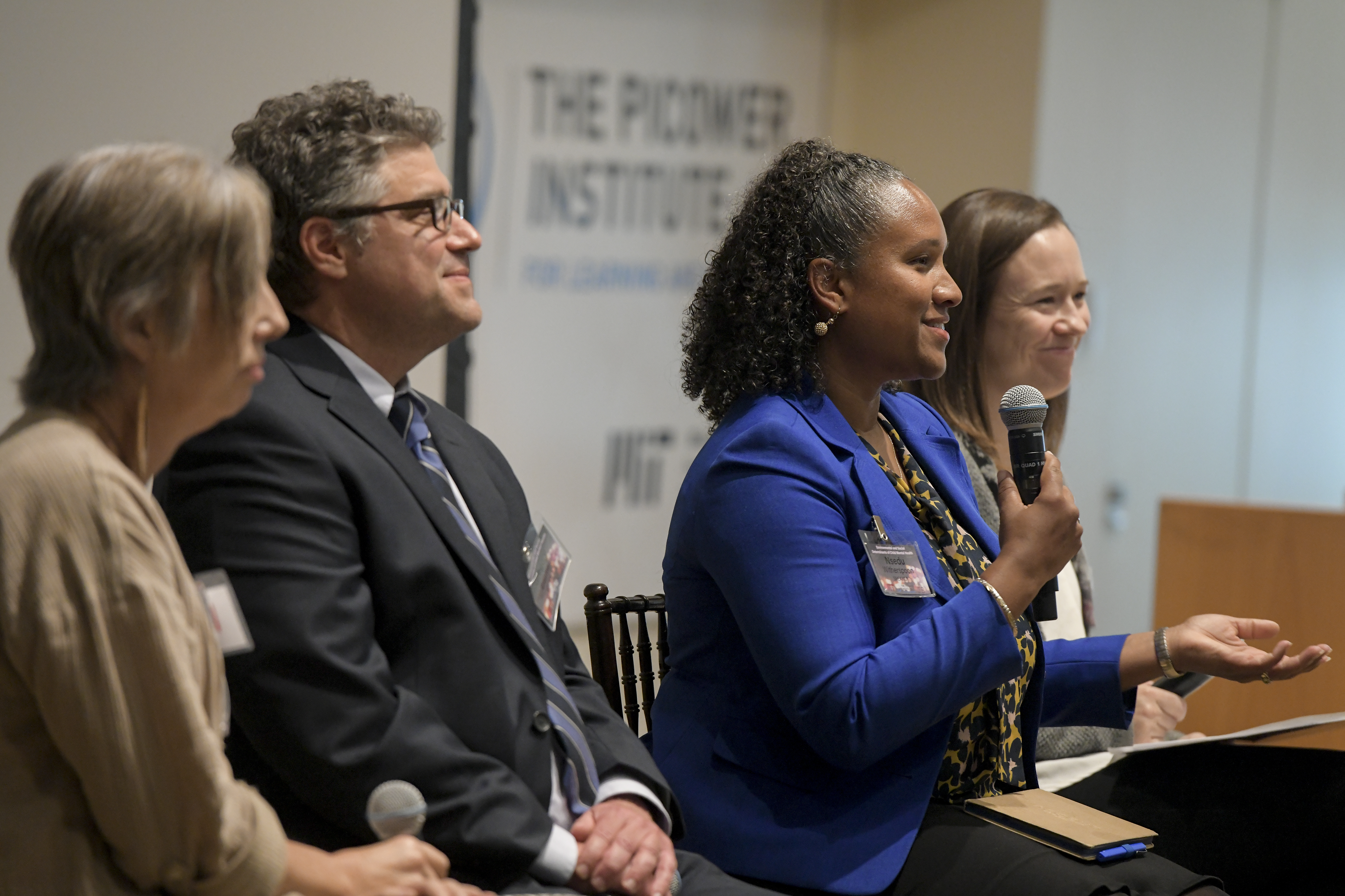 Four people -- three women and one man-- sit side by side. One woman. Nsedu Witherspoon, holds a microphone and is speaking.  The Picower Institute logo is in the background