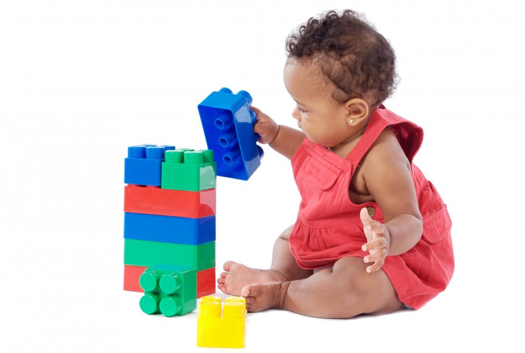 On a blank white background a baby girl sits and places a blue plastic brick upon a stack of other bricks of blue, green and red. Nearby a yellow block sits on the floor.