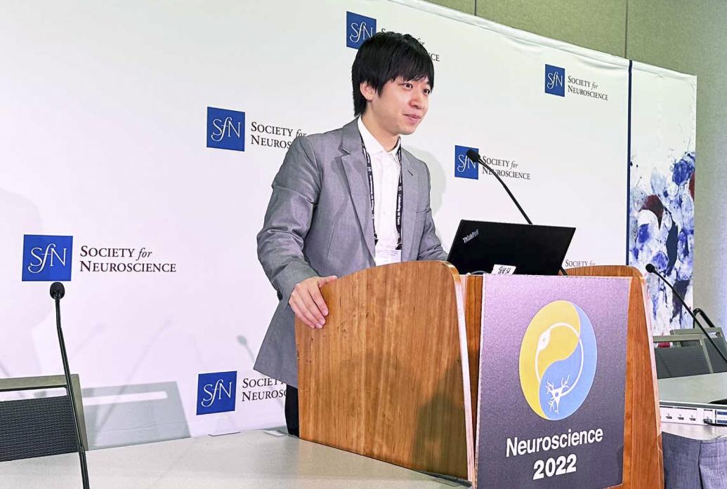 Takato Honda stands at a podium labeled Neuroscience 2022. Behind him is a white wall decorated with Society for Neuroscience logos