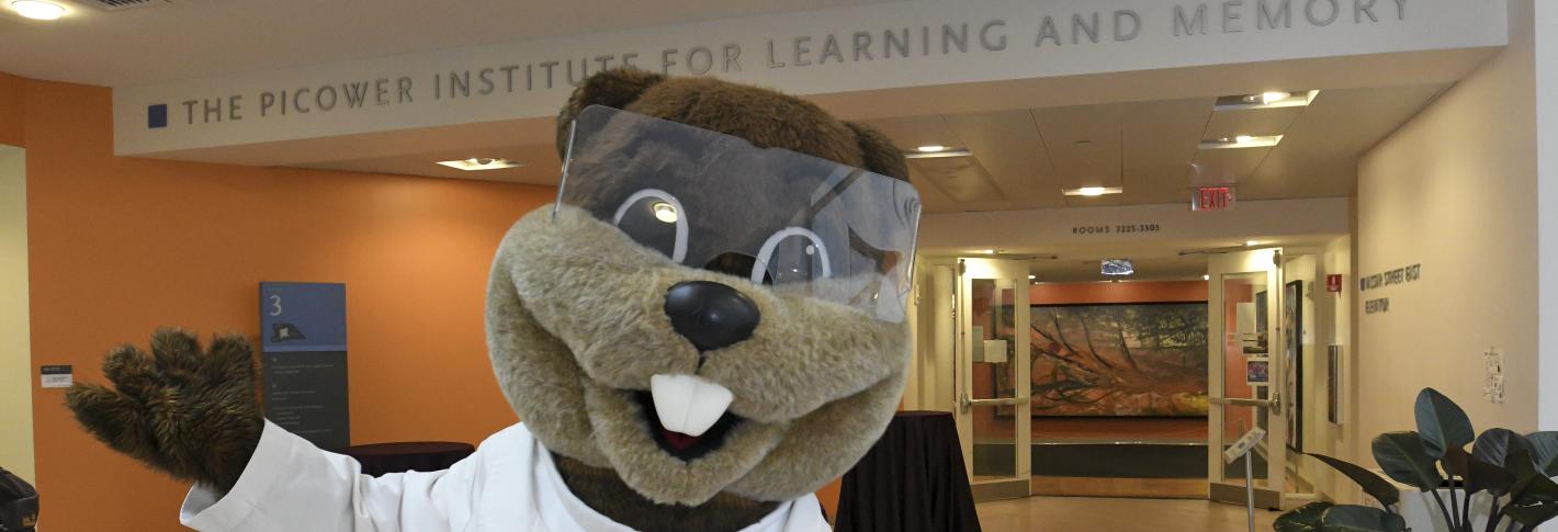 Tim the Beaver wears a lab coat. His arms extend joyously to his sides as he stands in front of the Picower Entrance off the Building 46 atrium