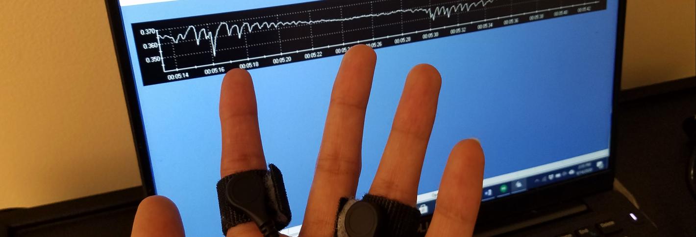 A hand with black bands strapped around the pointer and ringer is shown in front of a computer screen with a wiggly line stretching across