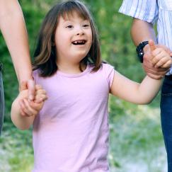 A girl with Down syndrome smiles while she holds her parents' hands.