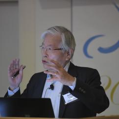 Susumu Tonegawa stands at a podium with the Picower Institute logo behind him. His hands are raised in an empatic gesture as he makes a point during a talk.
