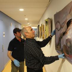 Todd Siler holds a painting up against a wall while an installer helps him assess its placement
