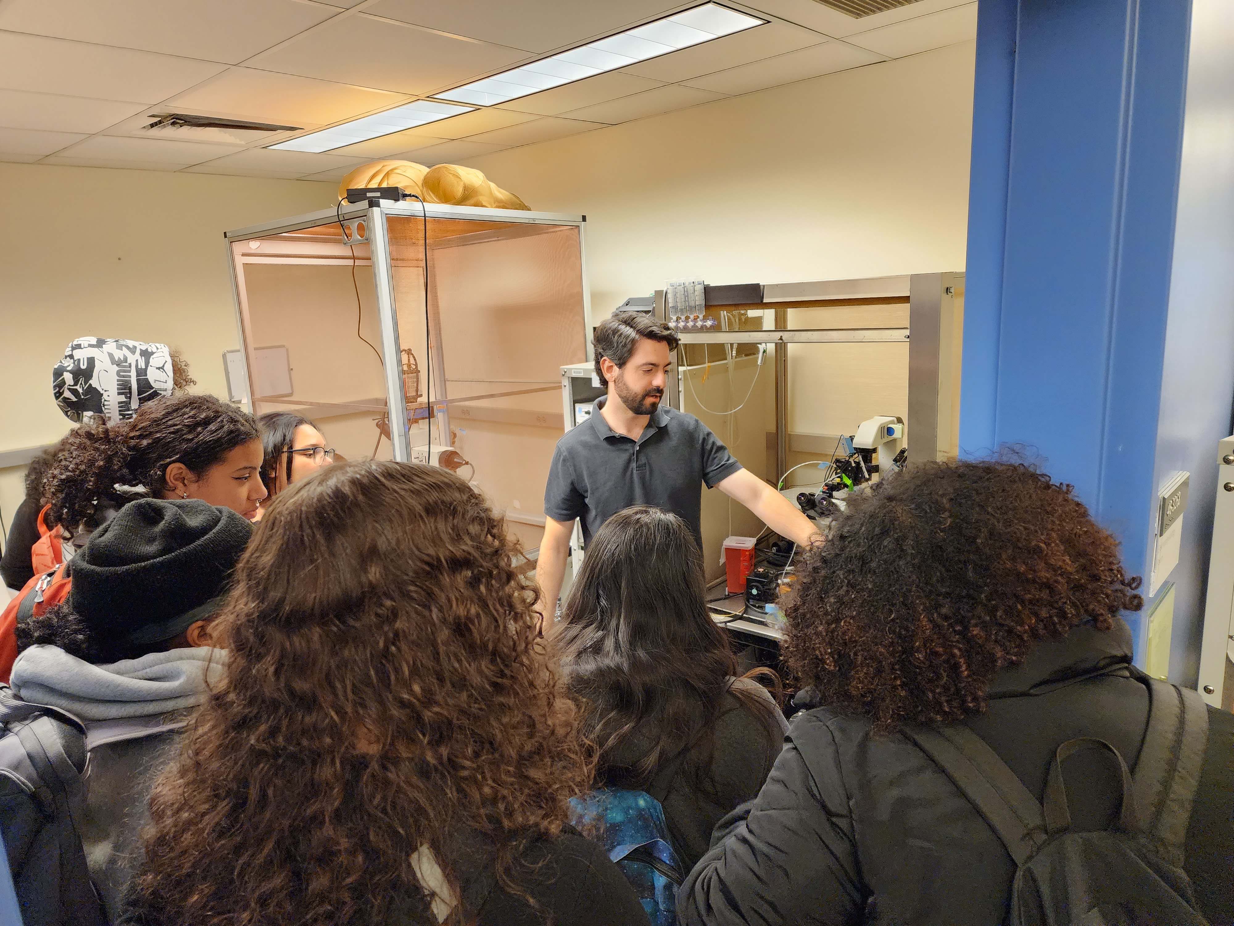 Andres Crane gestures toward an electrophysiology rig as seven high school students look on