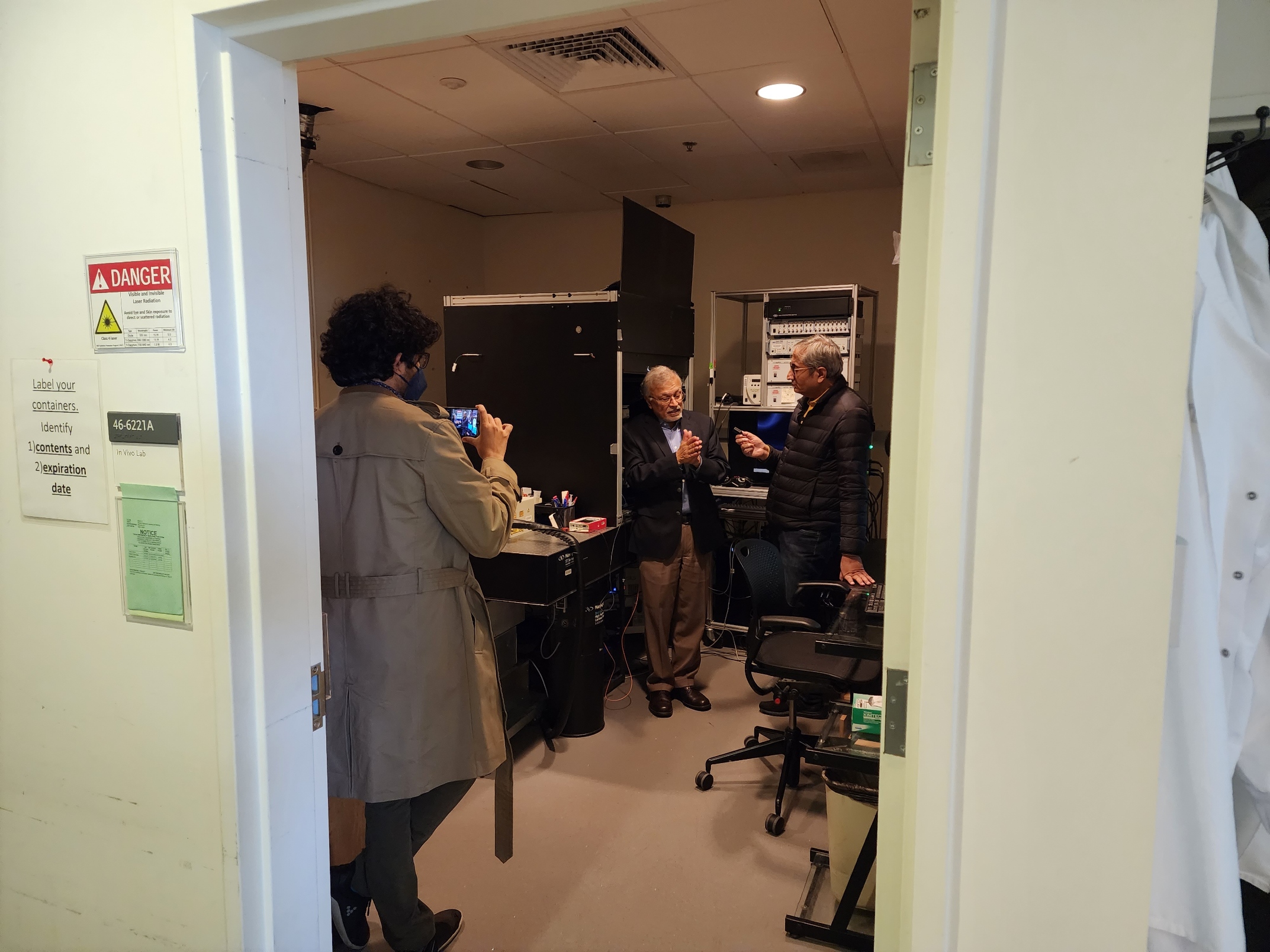 The camera peers through a doorway as a young man in a trenchcoat uses a phone to film Mriganka Sur and Ravish Kumar in conversation next to a large microscope and some computer equipment.