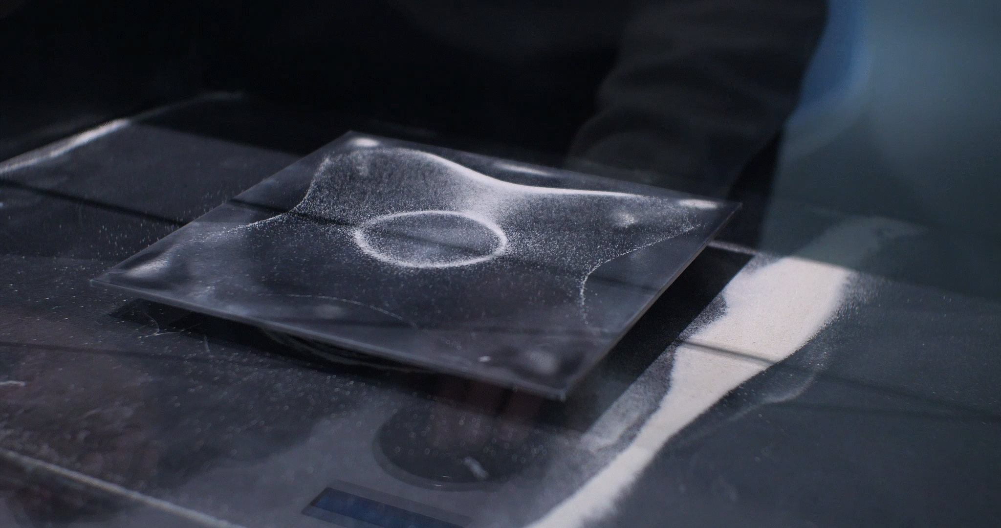 A platter set atop a speaker on a deejay's table holds grains of white sand. The sand grains align over the speaker, tracing out its underlying cloverleaf shape.