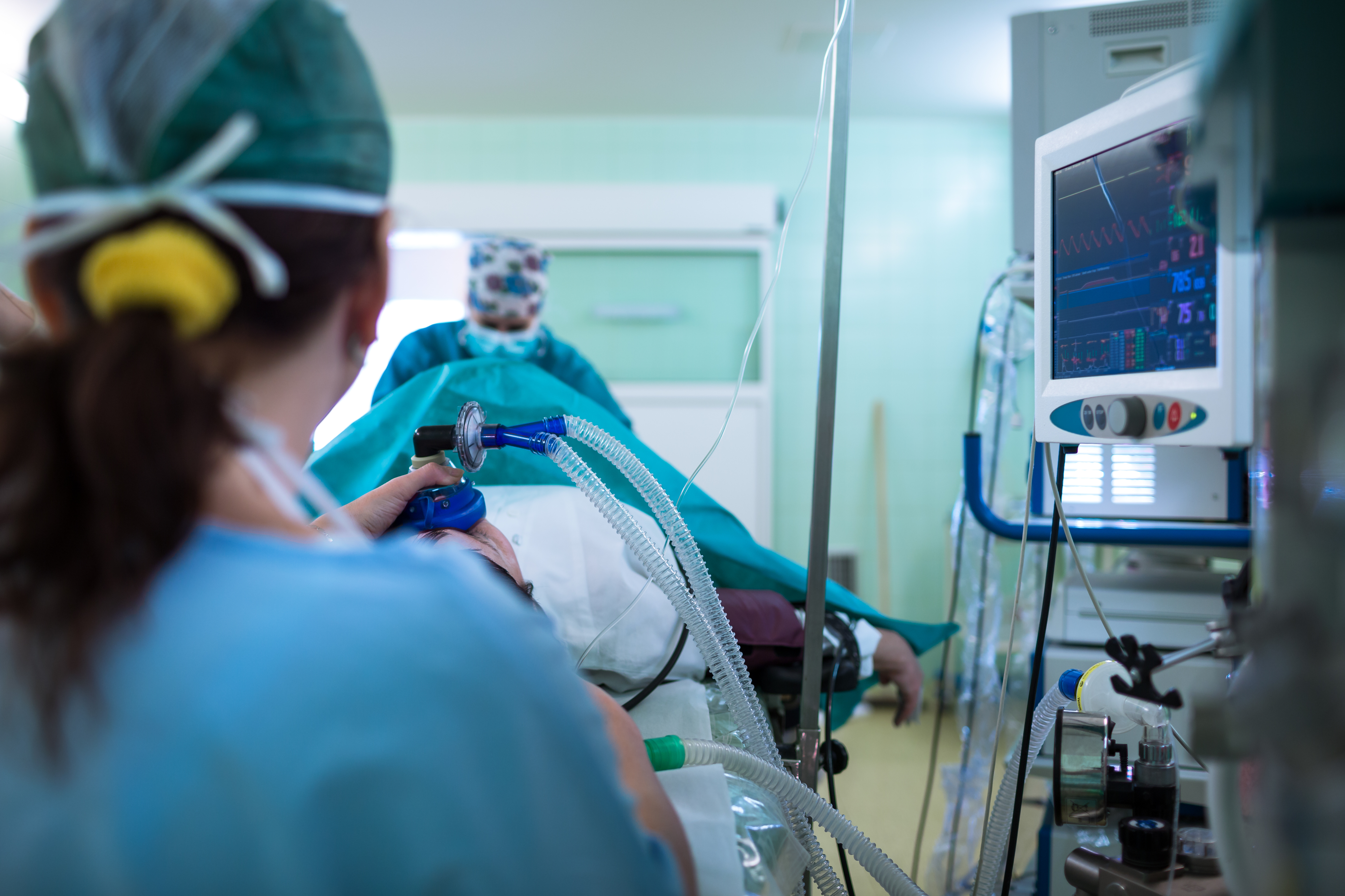An operating room scene shows a patient on a table. Our perspective is from behind the anesthesiologist who holds a mask on the patient's face and watches a monitor with a bunch of indicators. A surgeon stands out of focus on the far end of the patient.