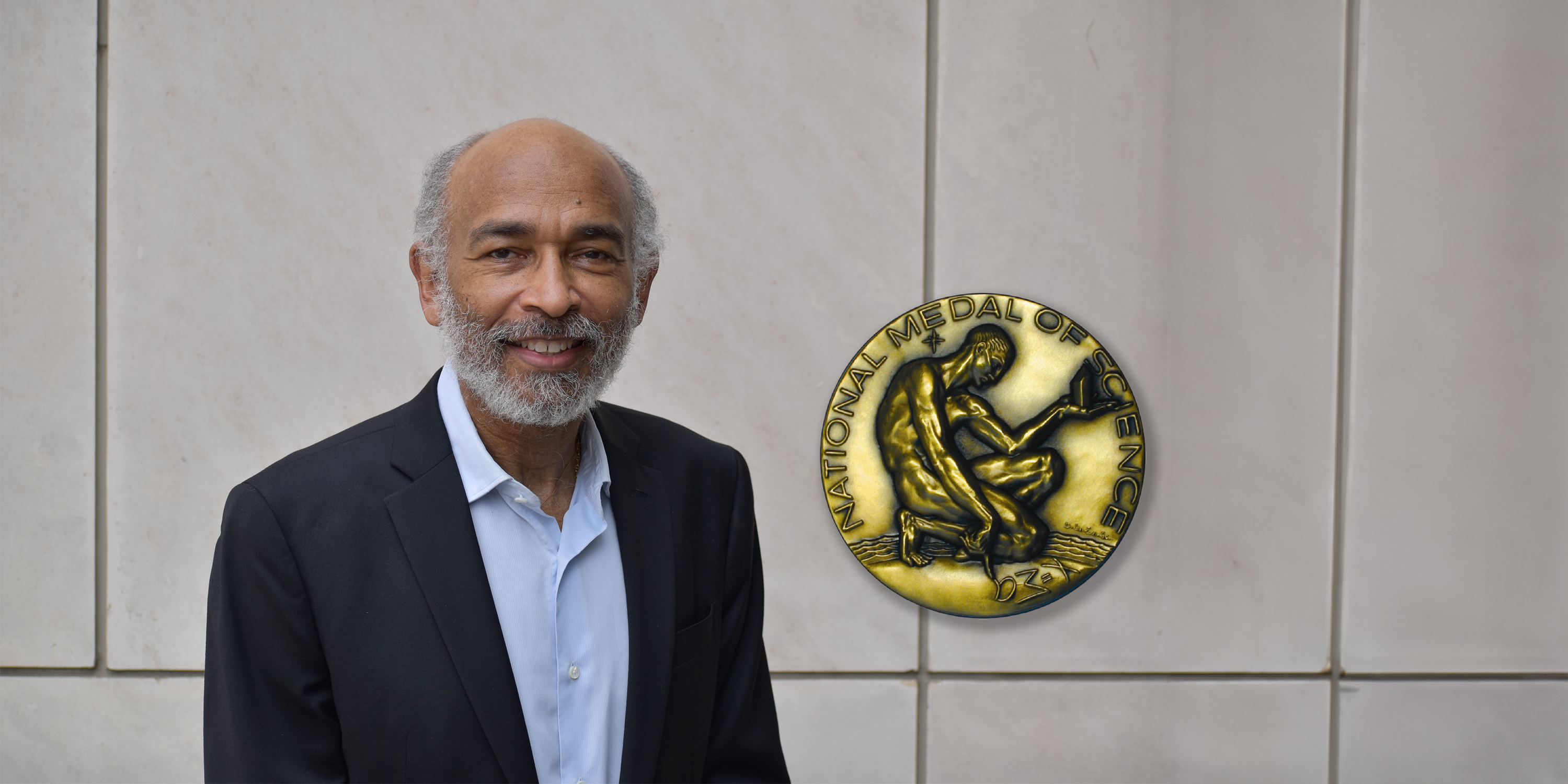 Emery N. Brown stands in front of a wall. Superimposed on the image next to him is a picture of the front of the National Medal of Science, which depicts a man kneeling and drawing an abstract character in the ground next to him as he contemplates an object.