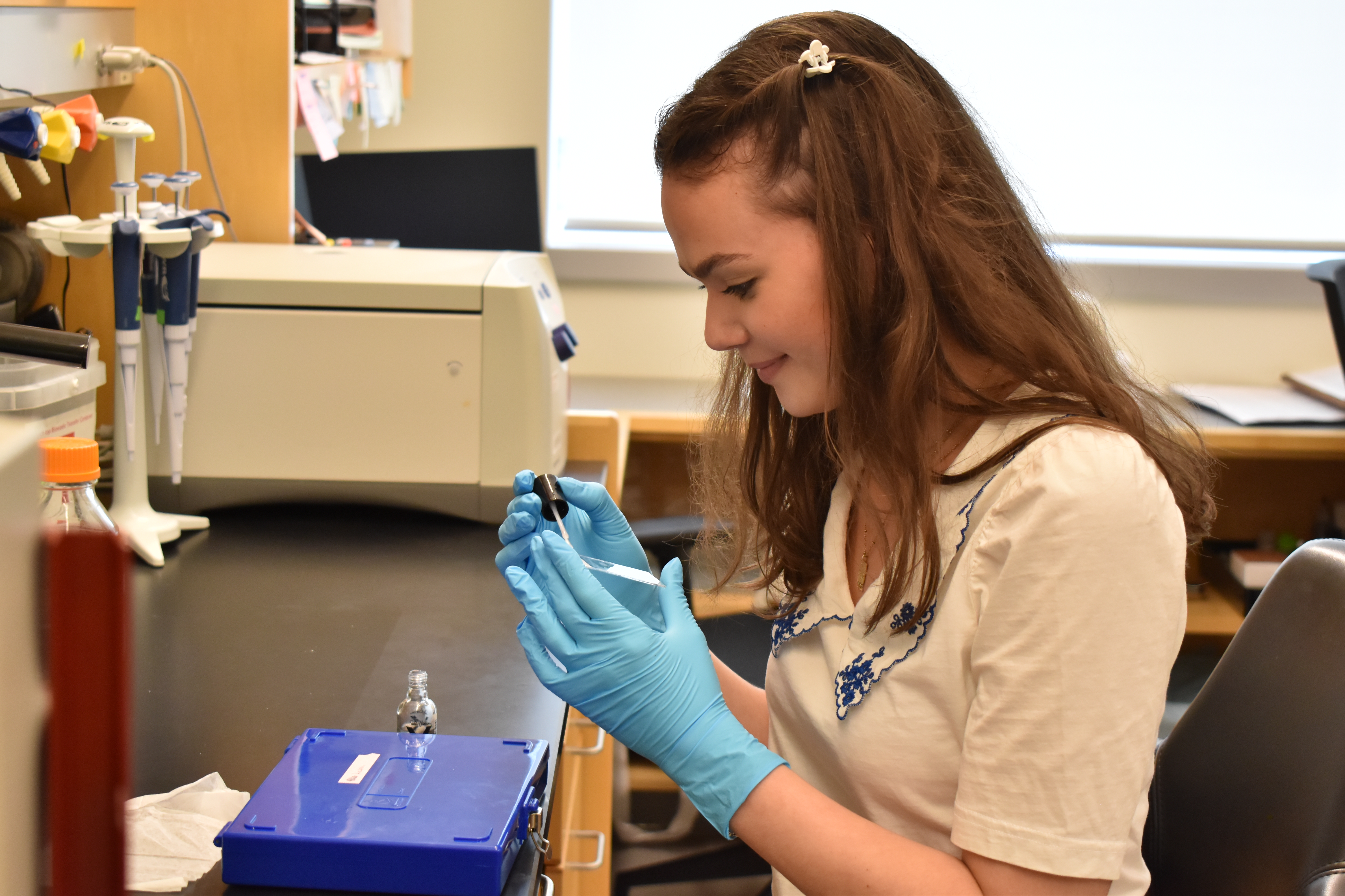 Laura Neal sits at a lab bench as she applies nail polish to the outer edges of the cover of a microscope slide. A blue plastic box of other slides is on the bench in front of her.