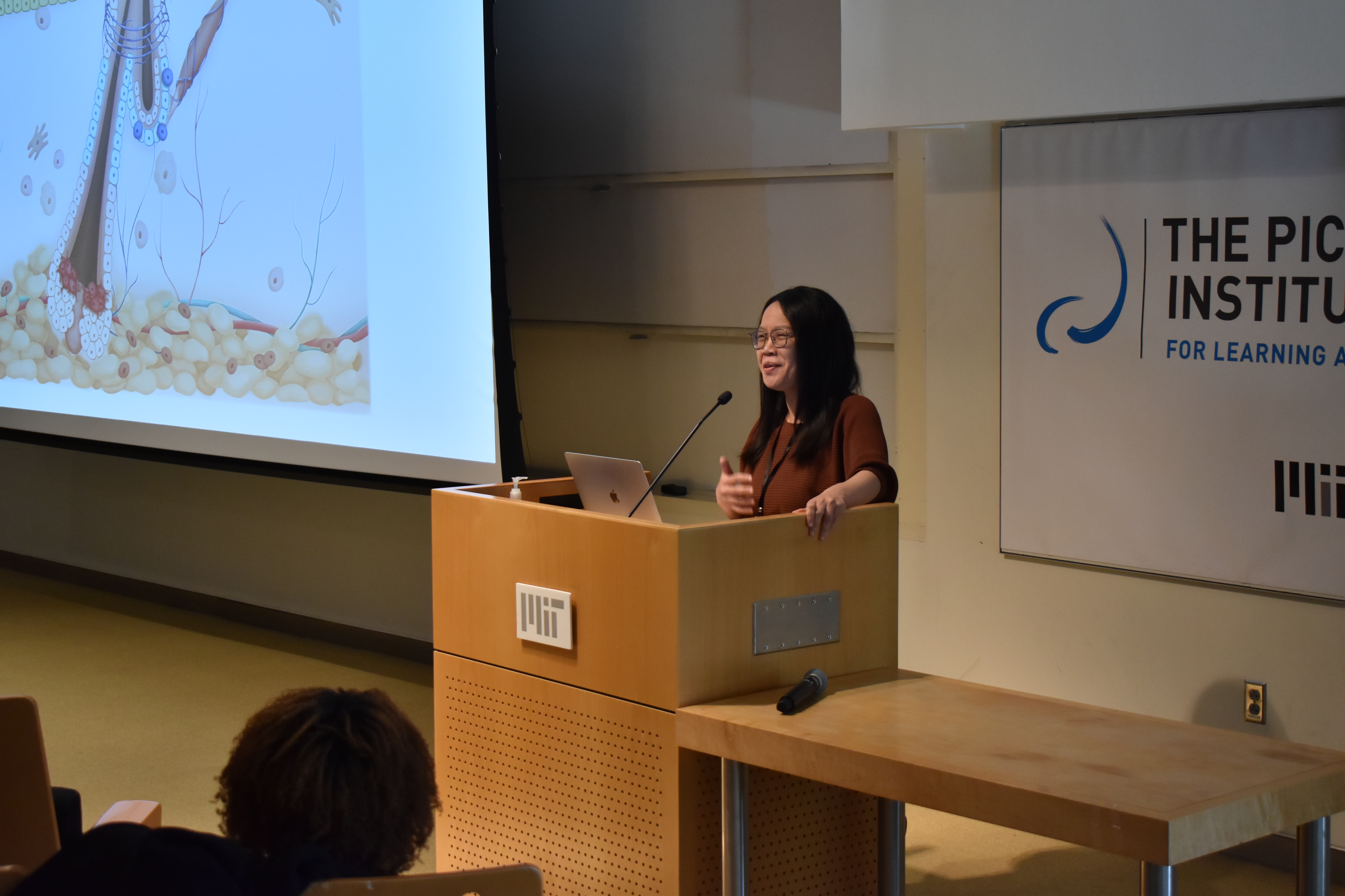 Ya-Chieh Hsu stands at a podium and speaks. On the screen to her right there is a large cartoon of a hair follicle.