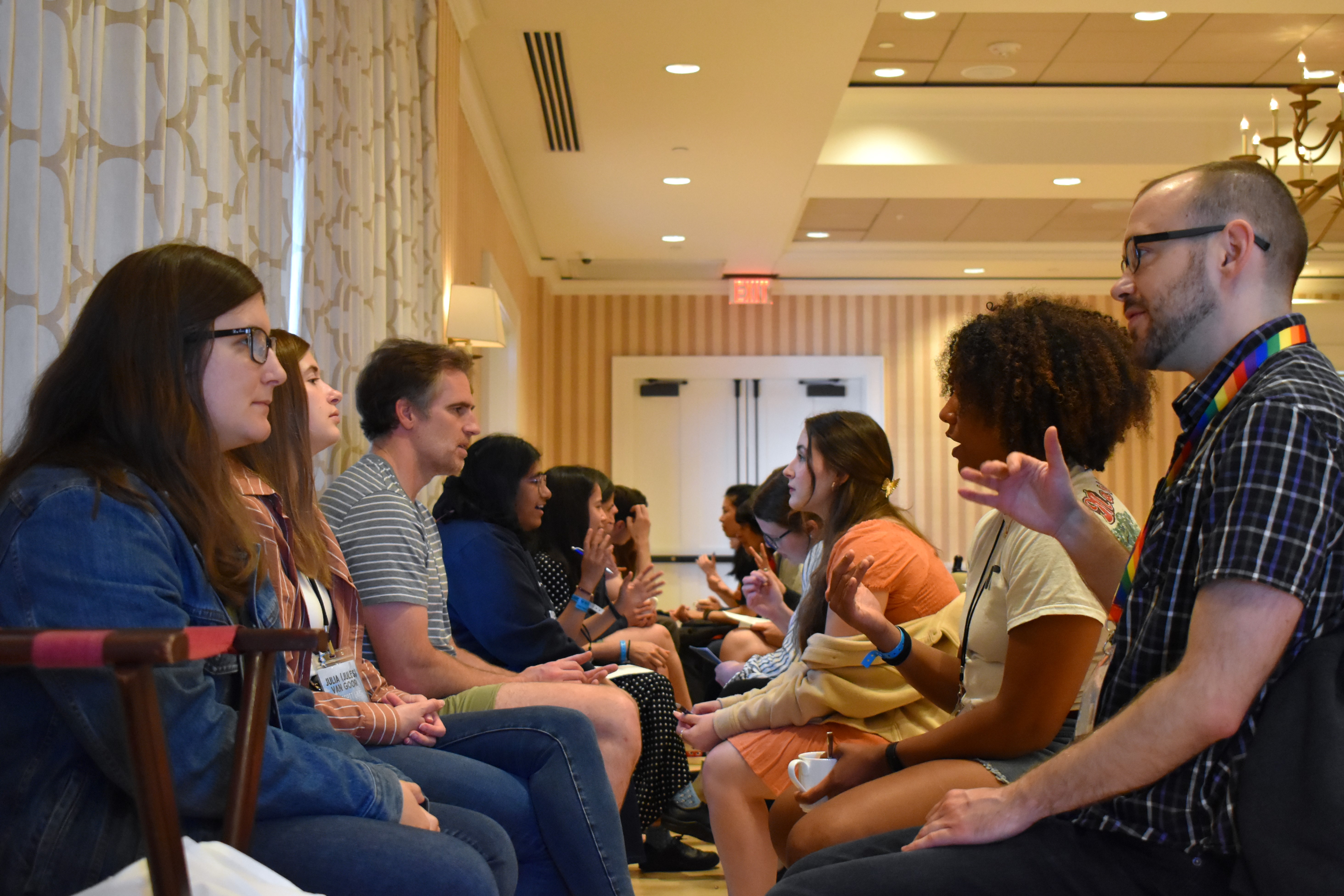 Two long rows of people sit across from eachother, facing eachother and conversing. The camera looks down the line between the rows.