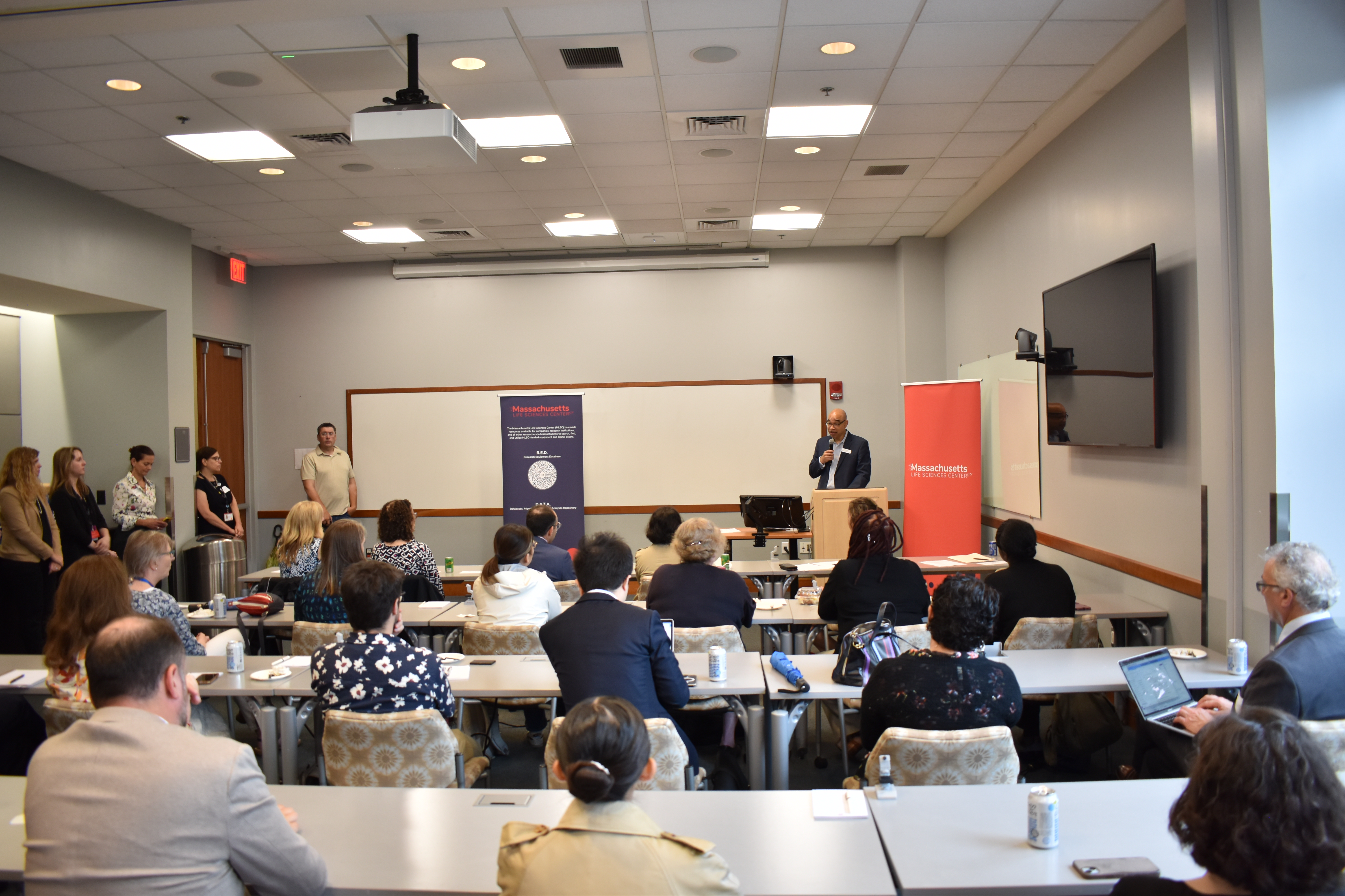 A man stands at a podium in front of a room full of more than 70 people