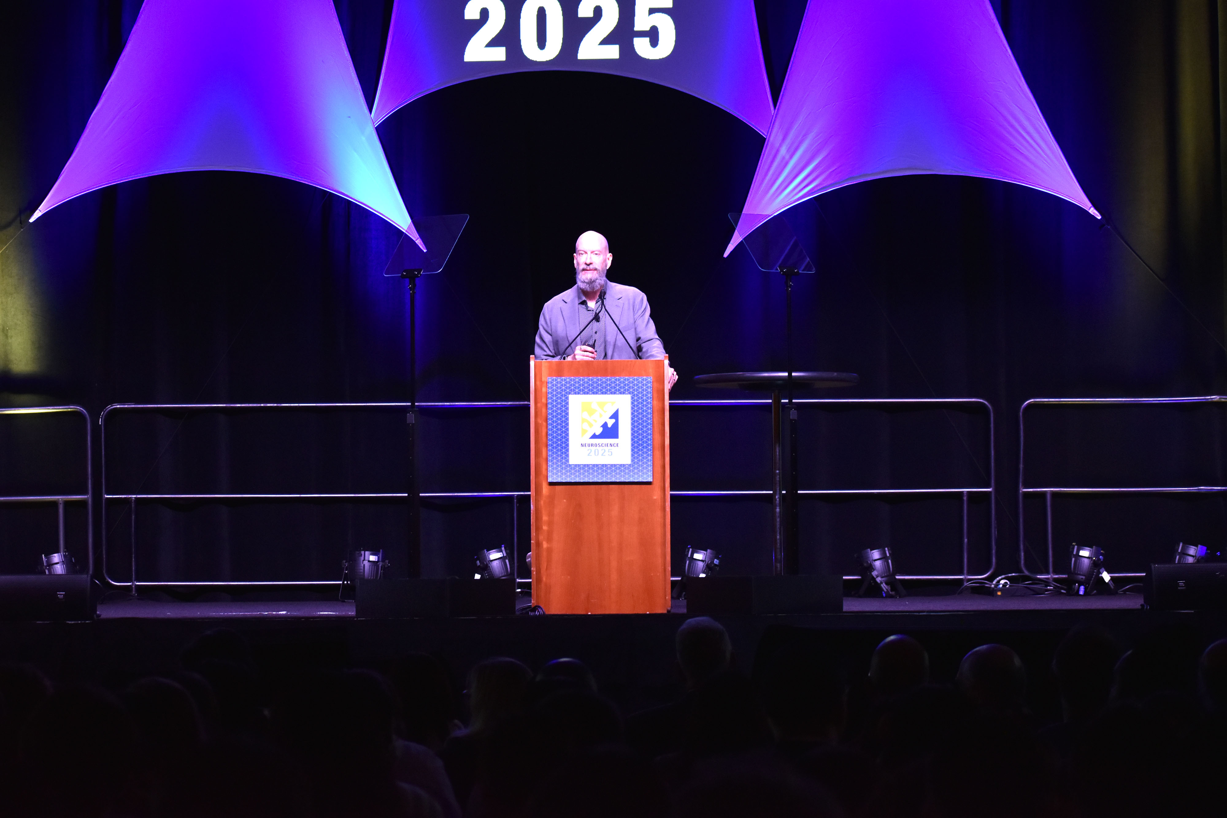 Earl Miller stands at the podium decorated with the Neuroscience 2025 logo and with audience members sitting in the foreground.