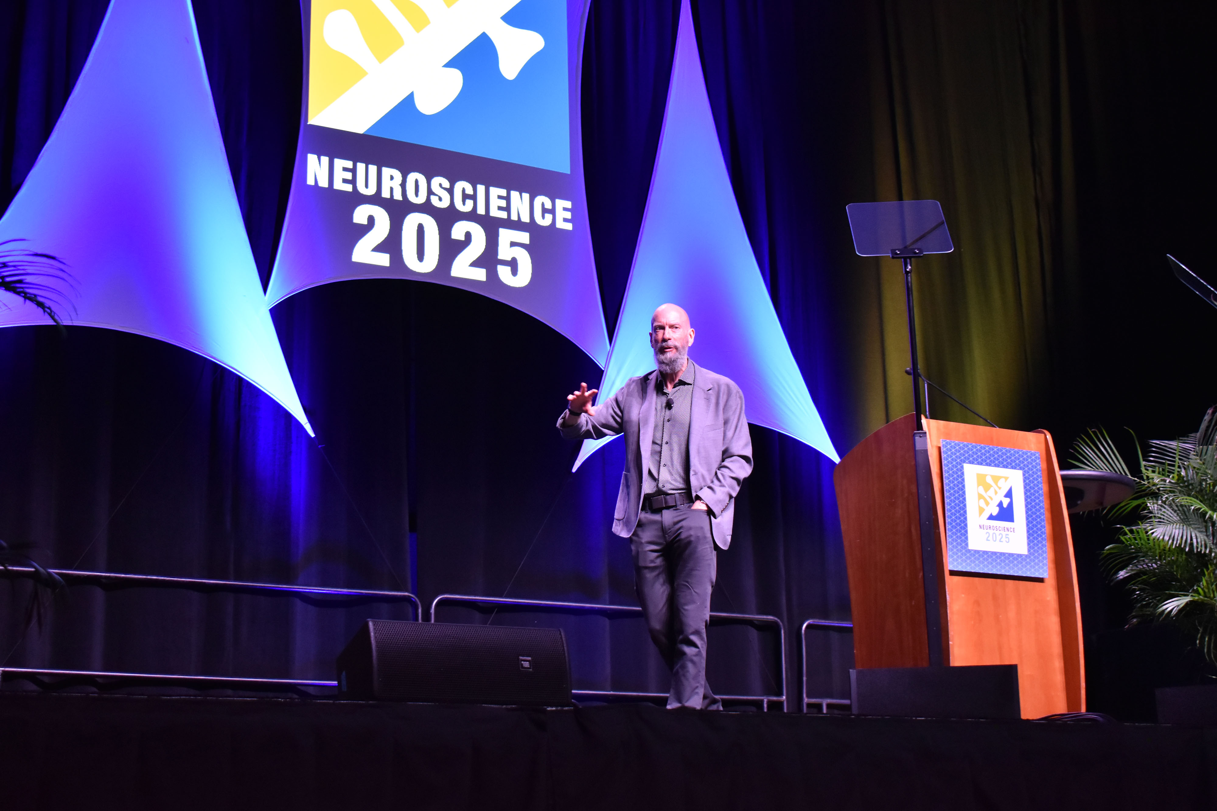 Earl Miller gestures walks across a large stage decorated with the words "Neuroscience 2025"