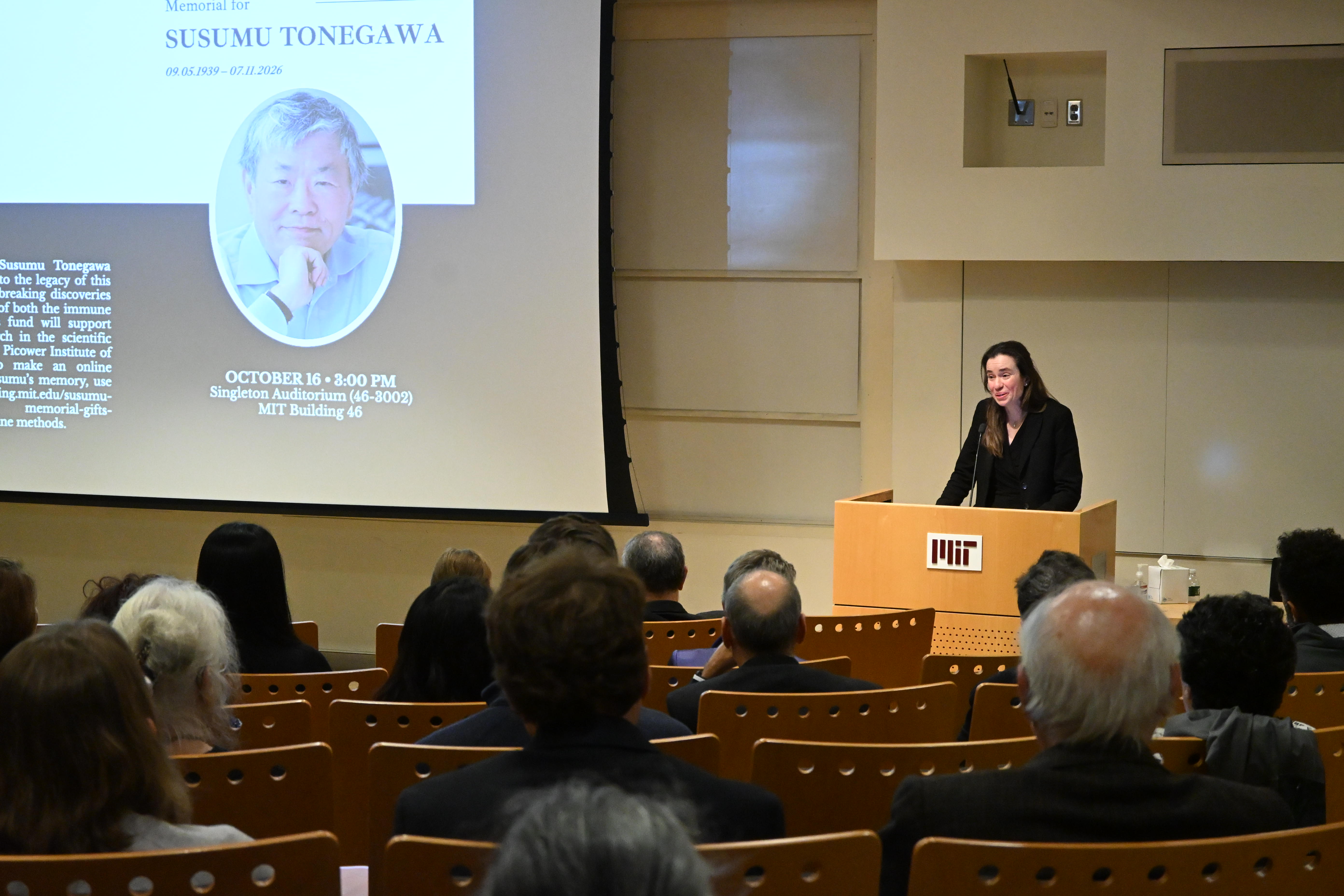 Myriam Heiman stands at the podium of Singleton Auditorium in MIT's Building 46 with a picture of Susumu Tonegawa on the large screen to her right. An audience listens attentively in the foreground.