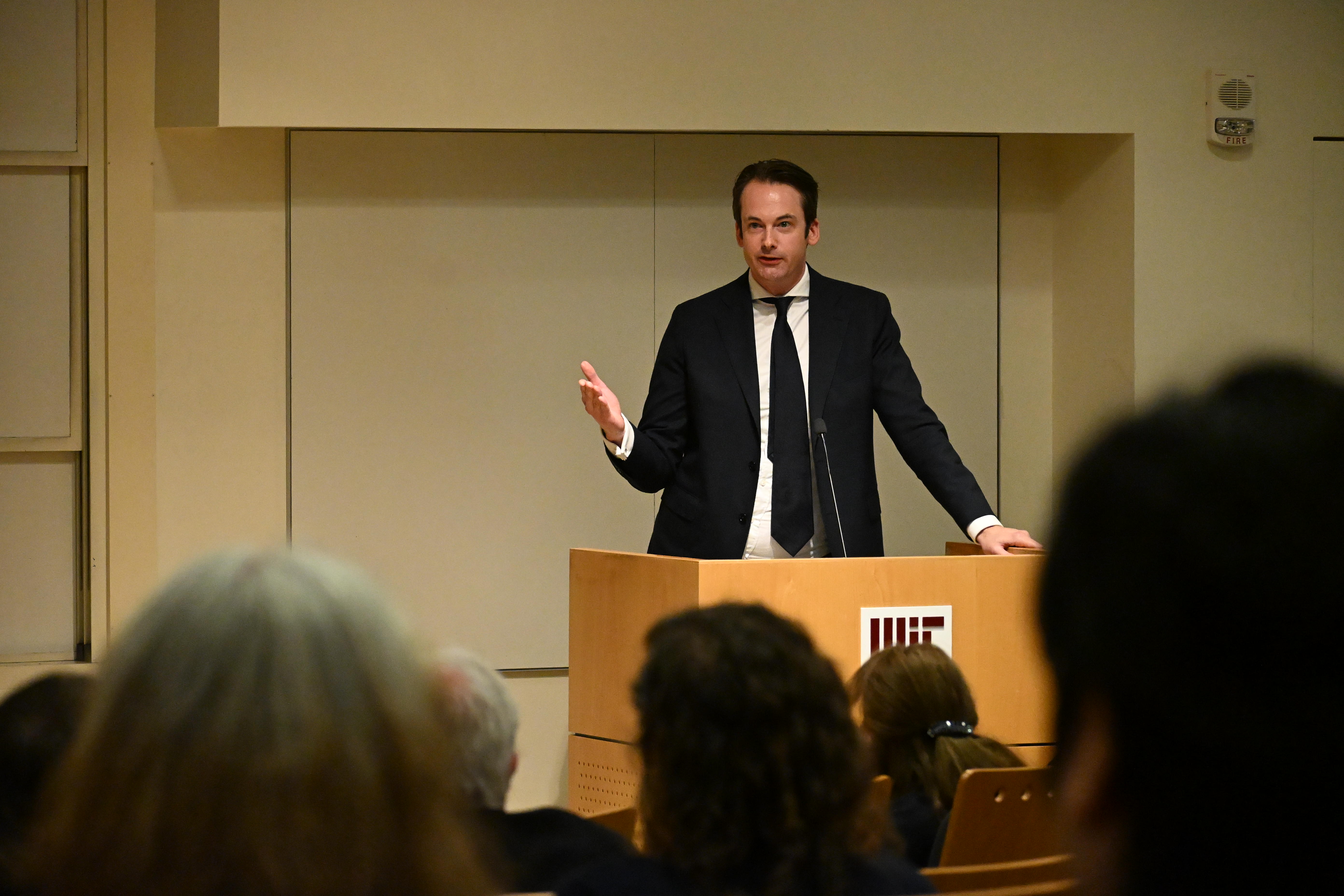 Tomas Ryan stands at the podium of Singleton Auditorium in MIT's Building 46 with the event audience in the foreground.