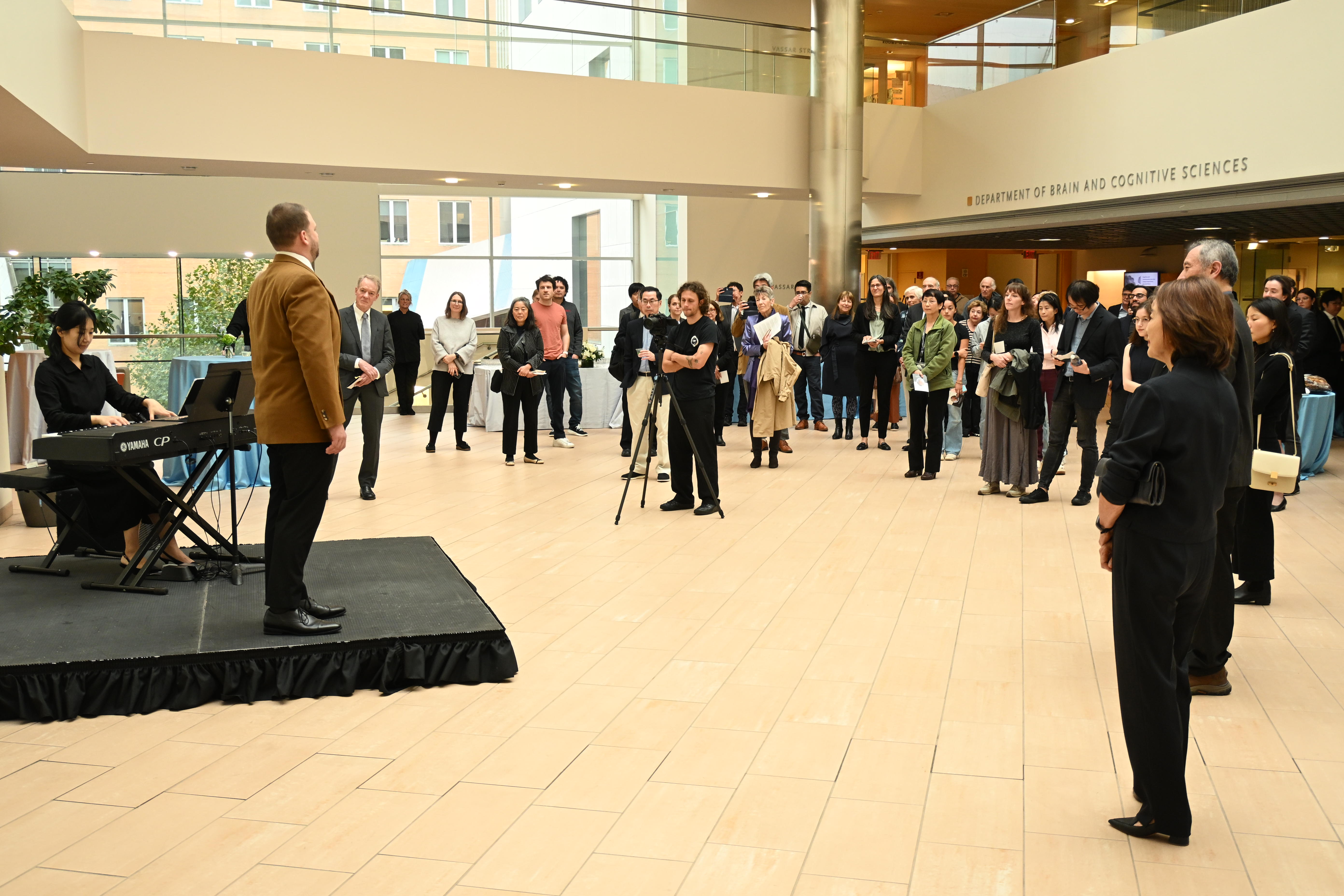 A crowd of people forms an arc around a small stage in the atrium of MIT's Building 46. On the stage is a woman playing a keyboard while a man sings.