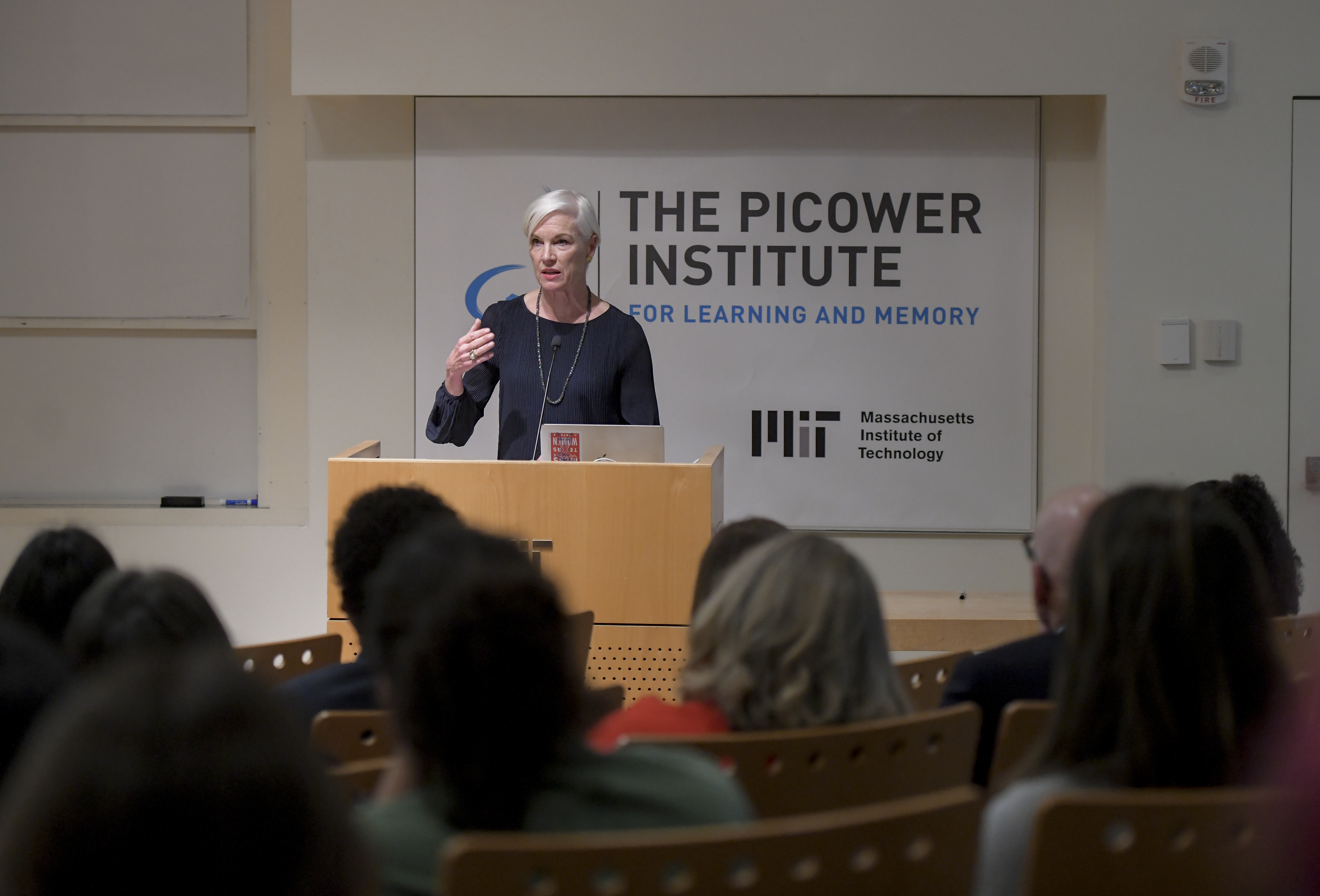 Cecile Richards stands behind a wooden podium in front of a background with the Picower Institute and MIT logos. In the foreground are several members of the audience out of focus.