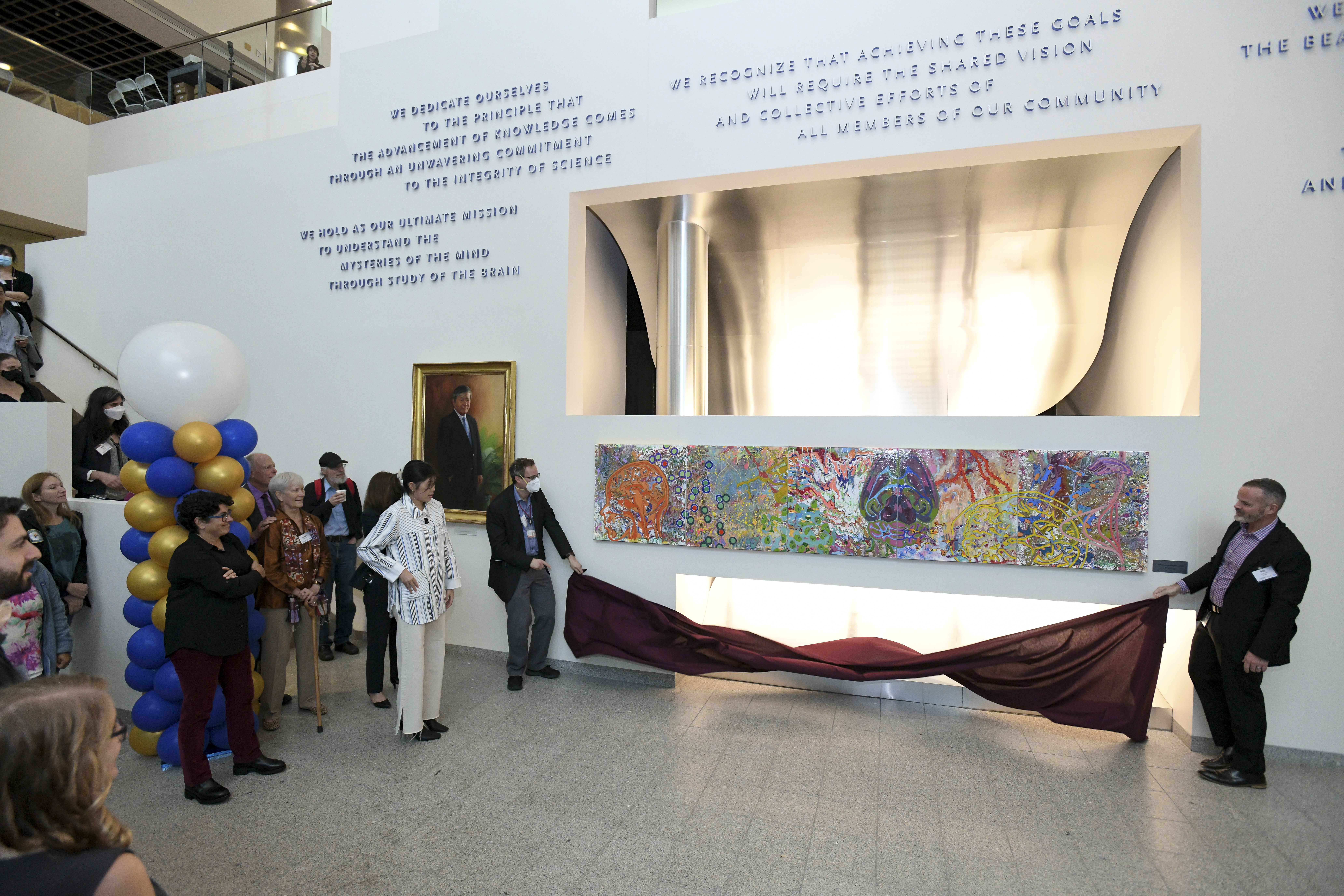 In the lobby of The Picower Institute in MIT's Building 46 two men hold a cloth under a wide, colorful painting composed of neuroscience imagery. Mila Sheng, the artist, stands nearby. To her right is Nergis Mavalvala. Standing behind them are many onlookers and a tall column of blue, gold and white balloons.