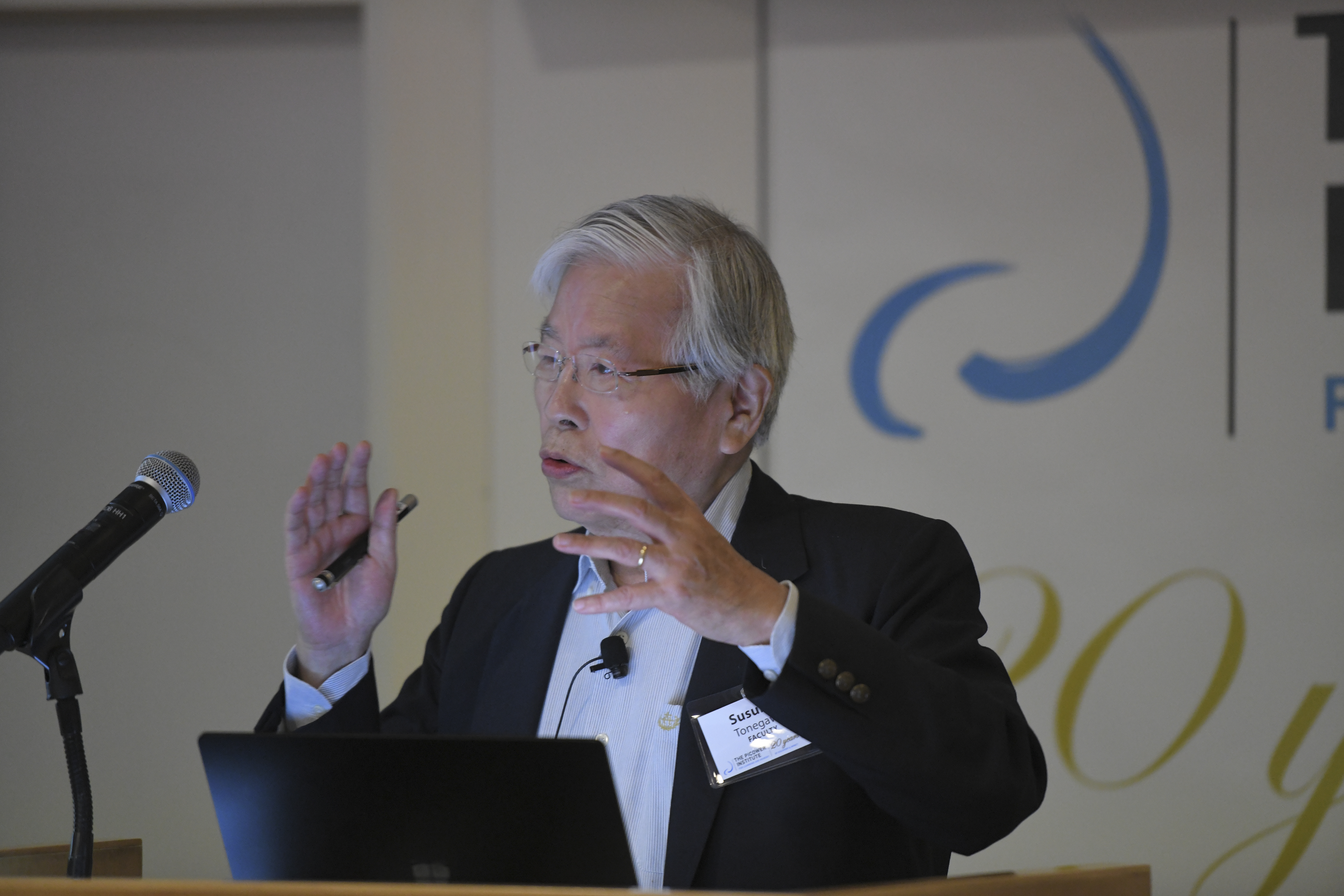 Susumu Tonegawa stands at a podium with the Picower Institute logo behind him. His hands are raised in an empatic gesture as he makes a point during a talk.