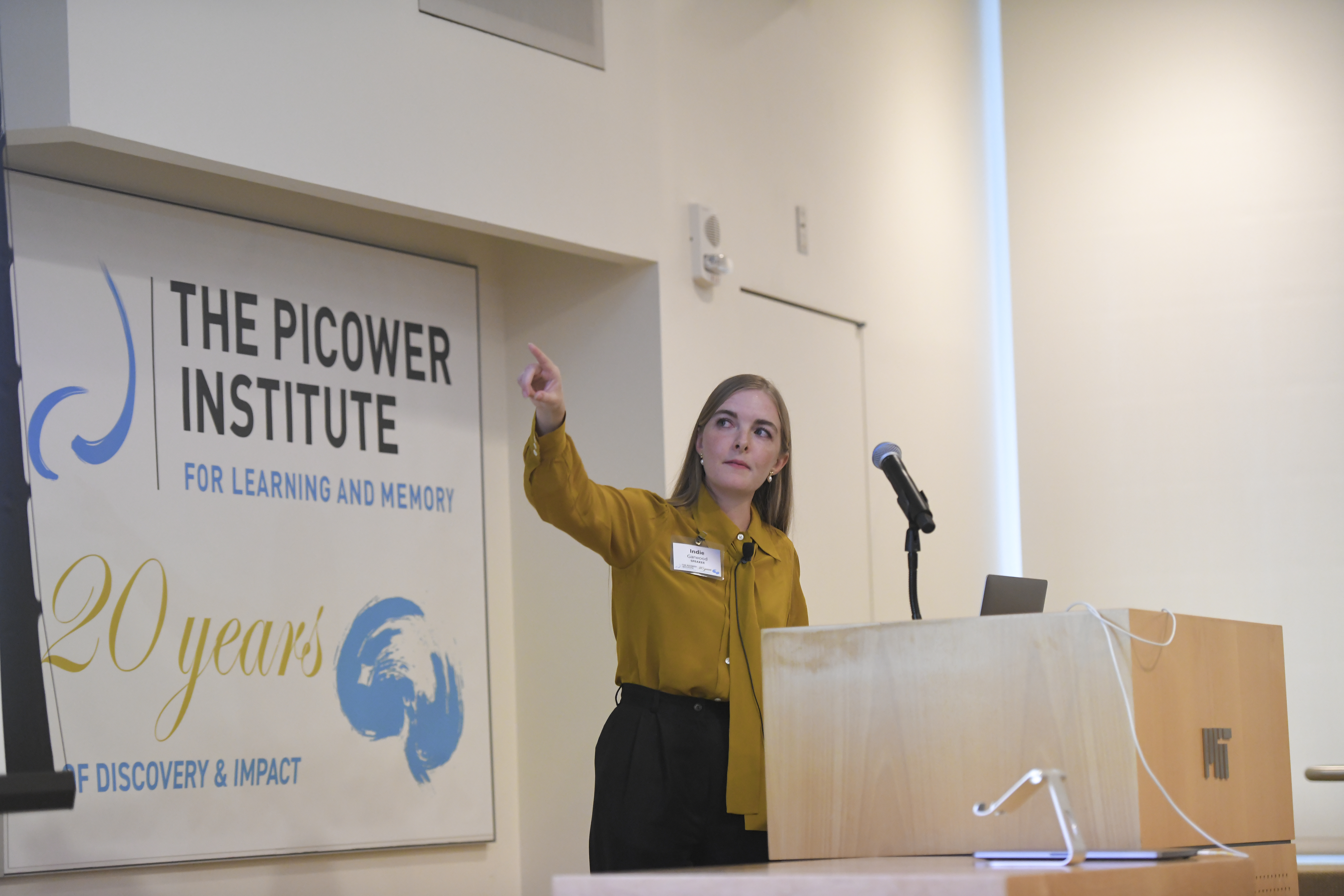 Indie Garwood stands at a podium during her presentation and points to her right (toward an unseen screen). Behind her is a sign showing the Picower Institute 20th Annivesary logo and 