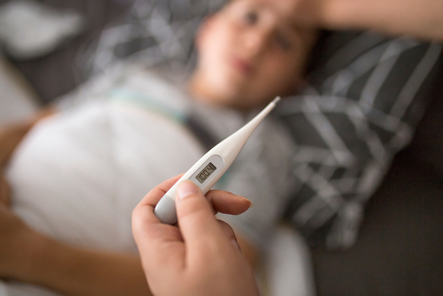 A thermometer shows a fever-level reading in the foreground as a sick child lies in the background