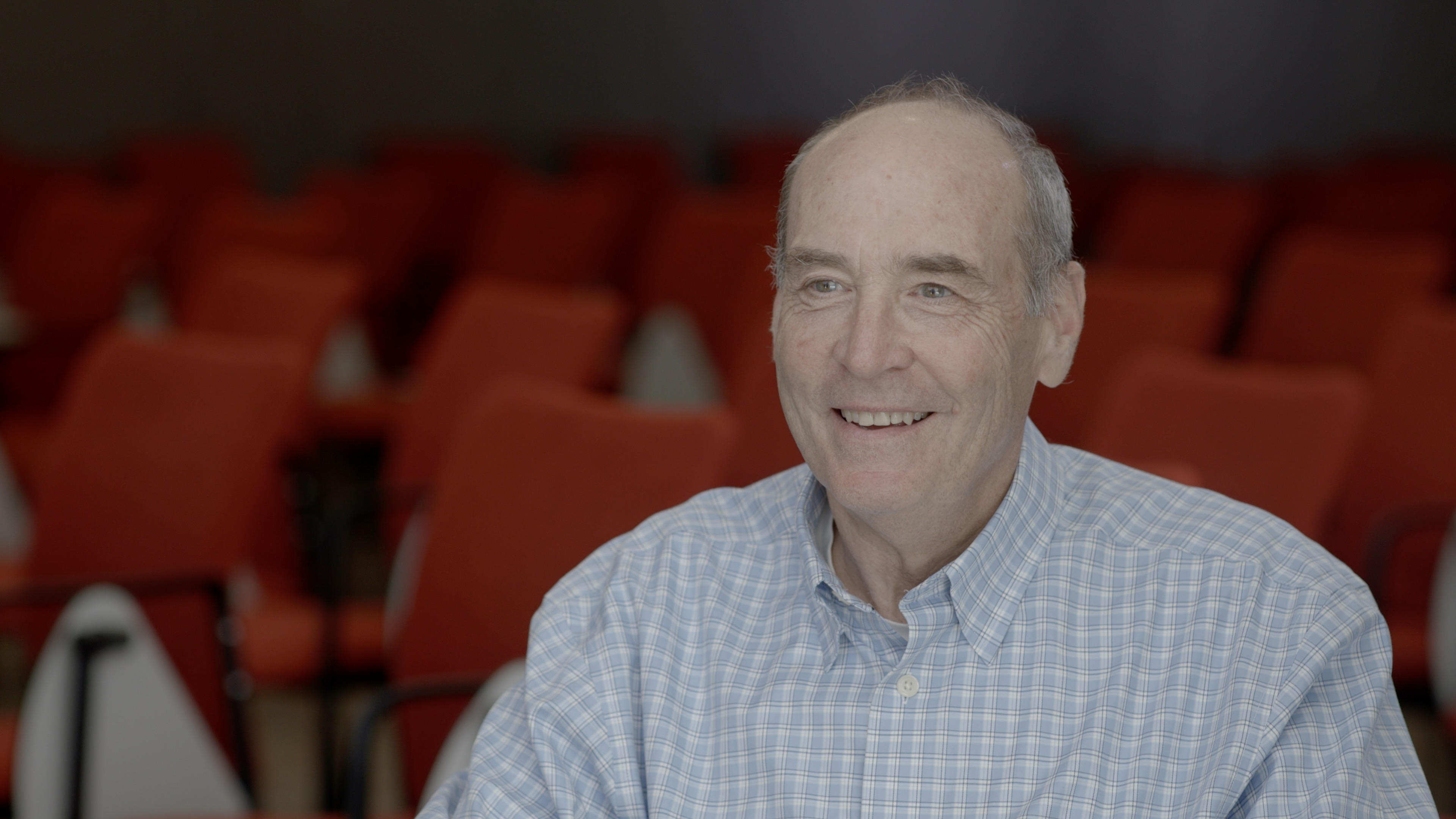 Mark Bear, shown from mid-chest up, smiles as he sits in a lecture room full of red chairs in the backgorund