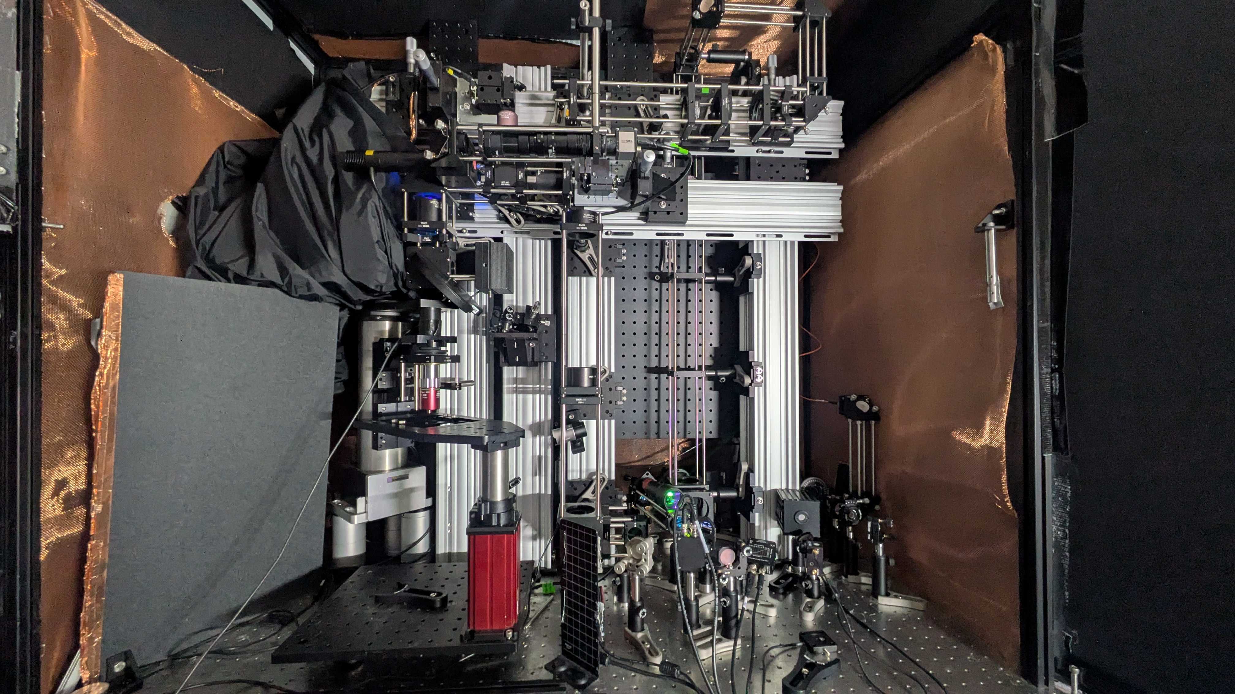 A microscope system, a complicated assemblage of optical, electronic and other equiptment built in several levels atop a metal table, is shielded around its side by dark paneling and copper colored screening.