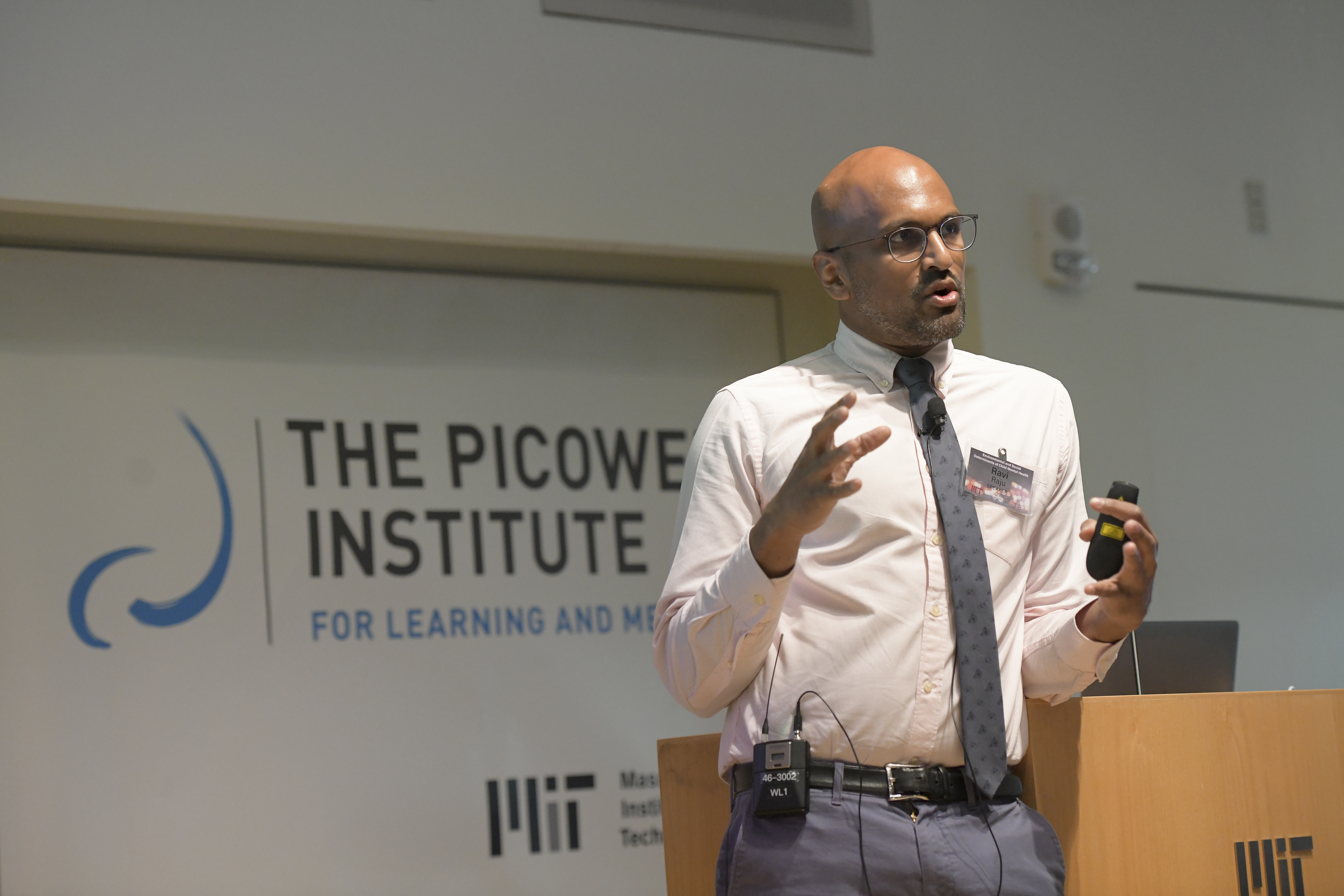 Ravi Raju, in a white shirt, tie and blue jeans speaks from a podium with the Picower Institute logo in the background