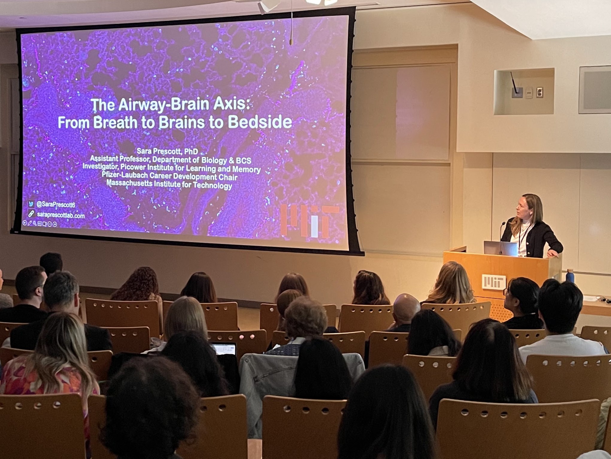 Sara Prescott stands at a podium in MIT's Building 46 delivering a talk to a large audience.