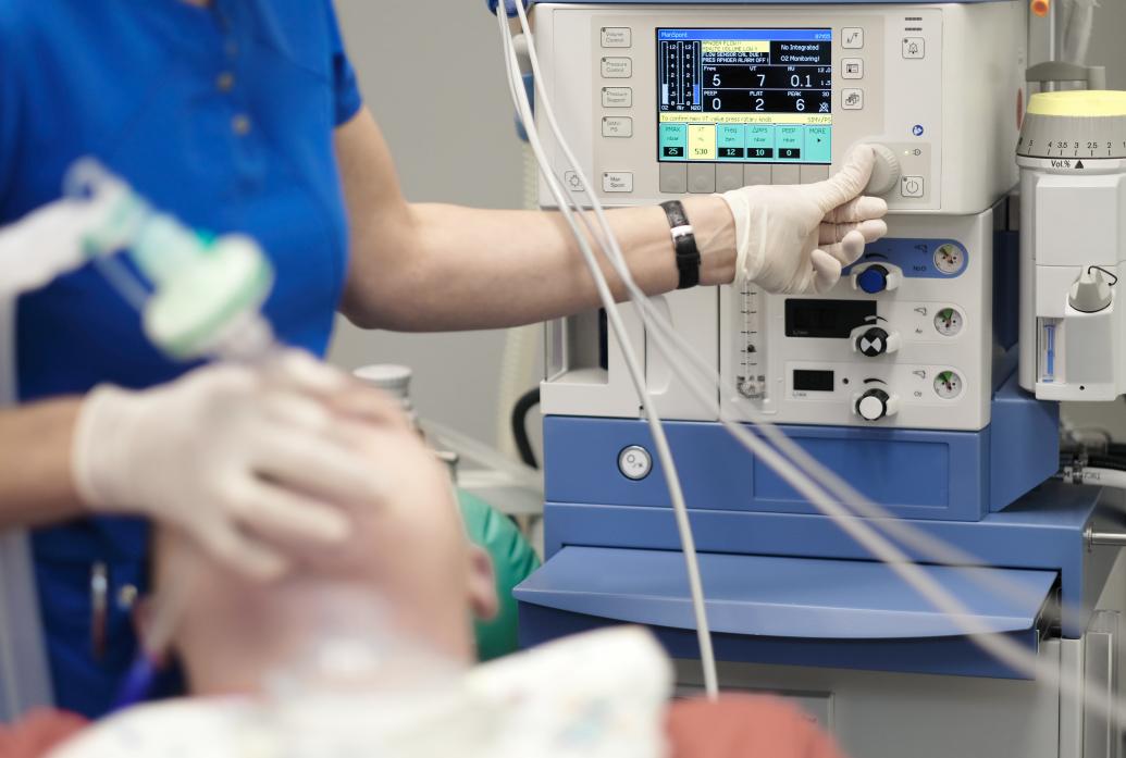 An anesthesiologist has his right hand on a patient's breathing tube and his left hand on a knob on a machine displaying various readings