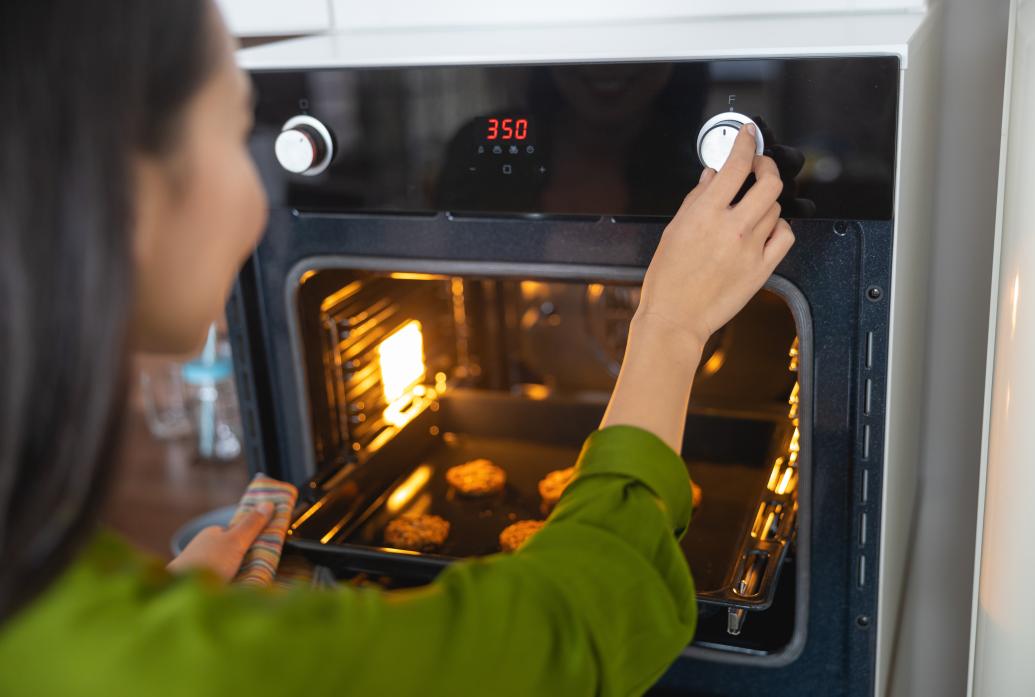 A woman turns a dial on an oven with cookies inside. The oven's display shows 350 indicating the temperature setting she has chosen.