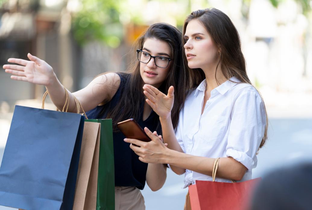 Two women on the street look intently and point in a particular direction. One holds a phone. The other holds shopping bags.
