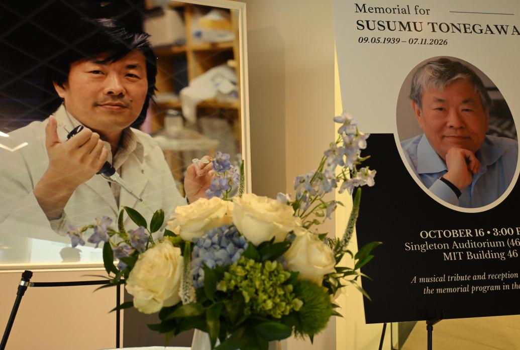 A bouquet of flowers sits in the foreground of two large photos on easels of Susumu Tonegawa at different stages of his career