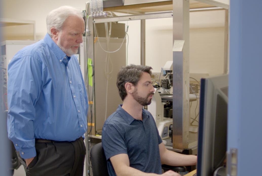 Andres Crane sits at a lab computer while Troy Littleton stands behind him looking over his shoulder at the screen.