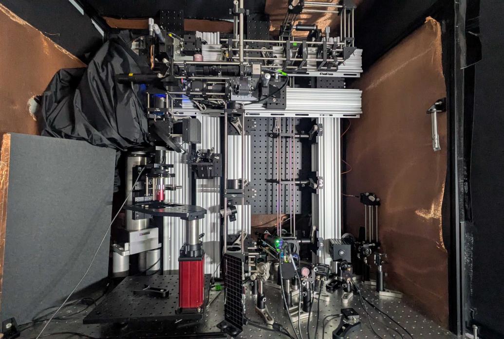 A microscope system, a complicated assemblage of optical, electronic and other equiptment built in several levels atop a metal table, is shielded around its side by dark paneling and copper colored screening.
