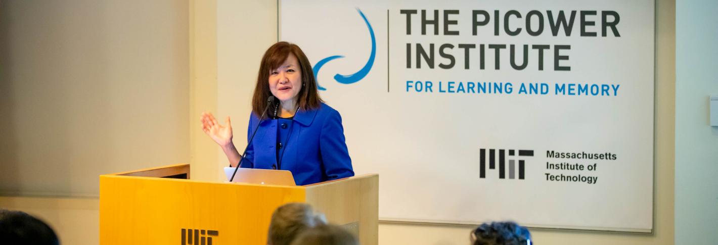 Li-Huei Tsai stands at a podium embossed with an MIT logo. In the foreground is an attentive audience. In the background is the Picower Institute logo
