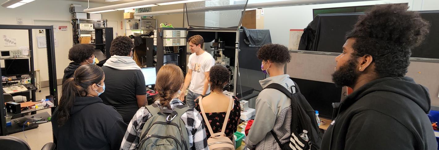 A group of students stands around a researcher in a white tee shirt as he explains the experiment he is performing with surrounding lab equipment.