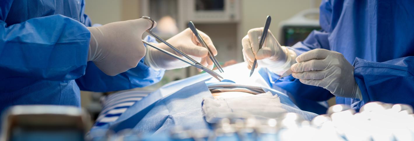 A view of a surgery in progress. Surgeons wearing blue gowns hold a scalpel and scissors just above an opening in the dressing of a patient who is lying on a table. Blurred in the foreground is a tray of other surgical instruments.