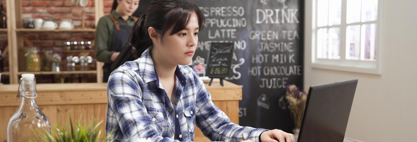 A young woman sits at a wooden table at a cafe staring at and typing on her laptop. The cafe counter is behind her and lots of menu options are written in chalk on a black menu board.