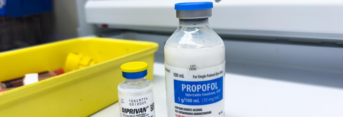 Two vials of propofol, a milky liquid, rest on a hospital shelf next to a small yelllow bin 