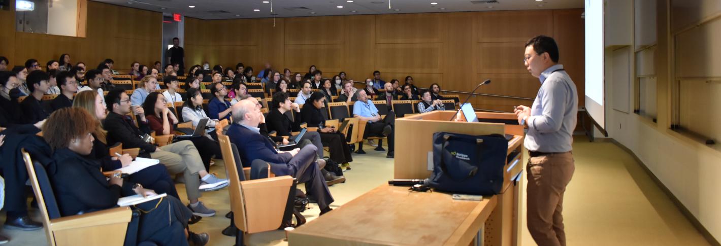 Li Ye is seen from the side as he delivers his talk at a symposium in MIT's Singleton Auditorium. In front of him is a nearly full room of audience members listening attentively.