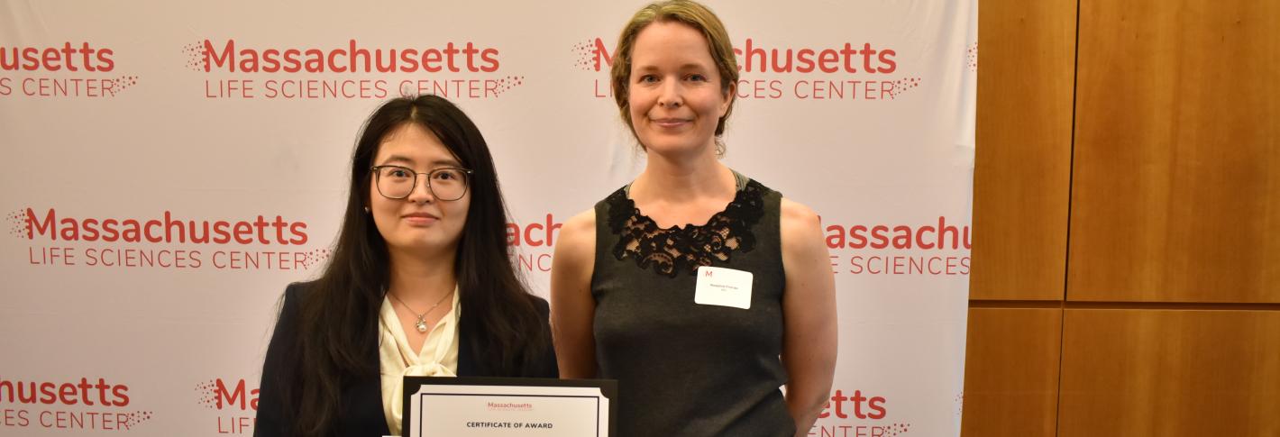 Shanshan Zhang and Rosie Firenze stand before a Massachusetts Life Sciences Center backdrop holding and award certificate