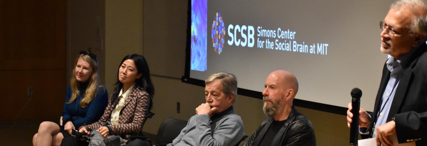 Ev Fedorenko, Gloria Choi, Charles Nelson and Earl Miller sit side by side on chairs in front of a screen displaying the logo of the Simons Center for the Social Brain.  Mriganka Sur stands nearby leaning on a podium and holding a microphone.