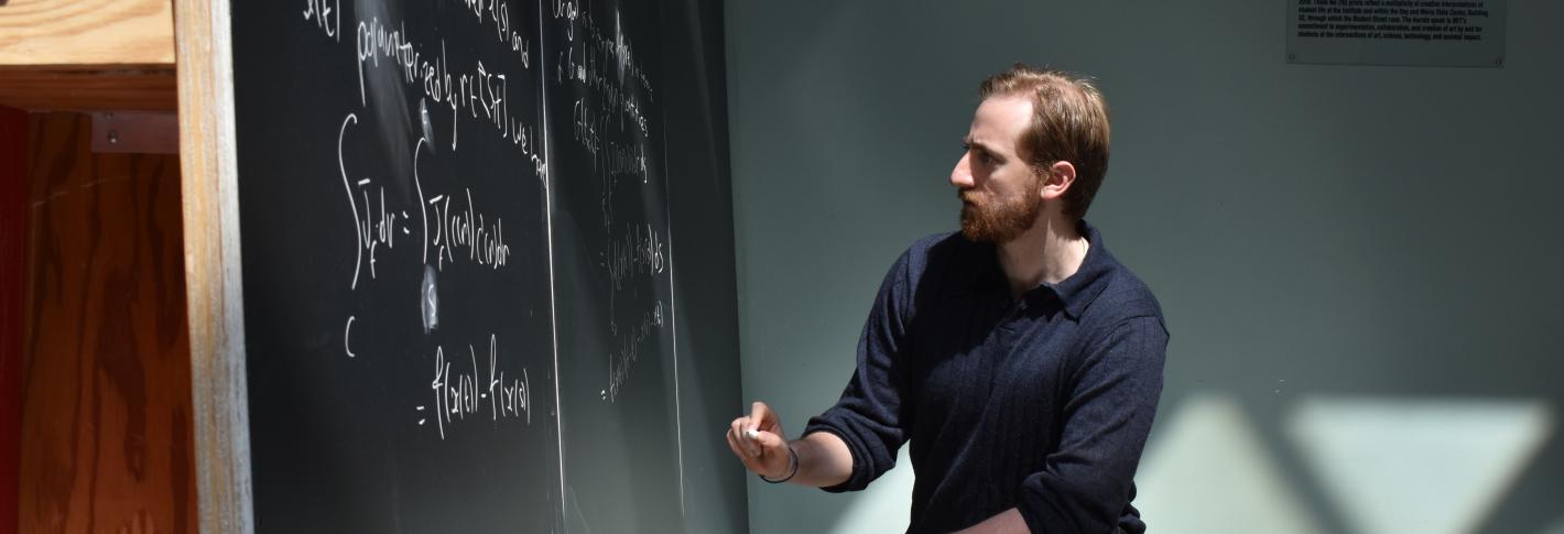Adam Eisen kneels and holds chalk, gazing intently at a large floor-to-ceiling chalkboard full of equations.