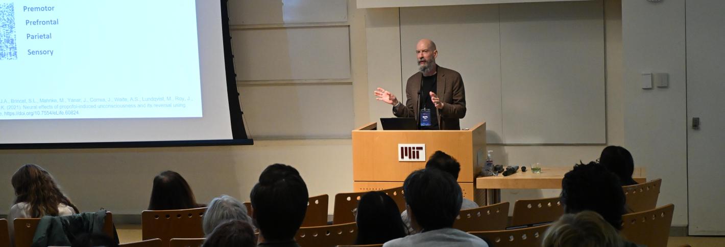 Earl Miller stands at a podium in MIT's Building 46, gesturing as he speaks to a room full of people.