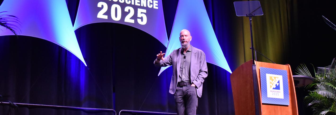 Earl Miller gestures walks across a large stage decorated with the words "Neuroscience 2025"