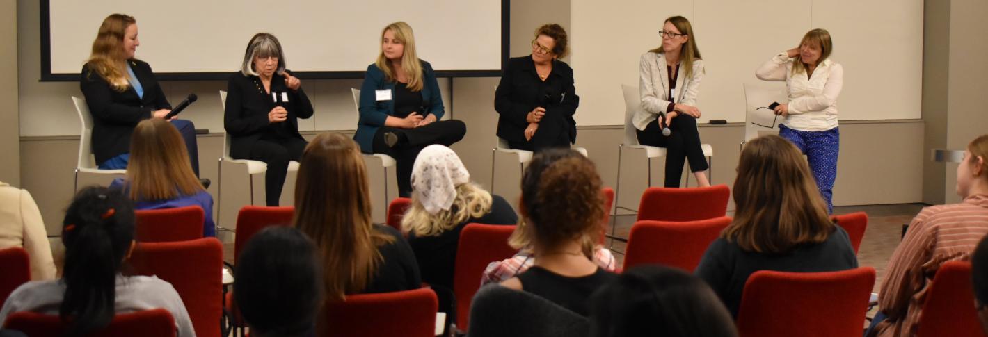 Six women, many holding microphones, sit in a row facing an audience of people sitting in red chairs.